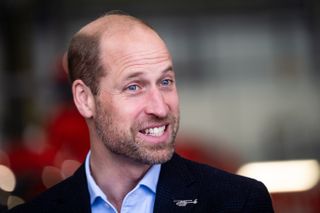 Prince William wearing a blue shirt and dark blazer with a helicopter pin and smiling looking to the right