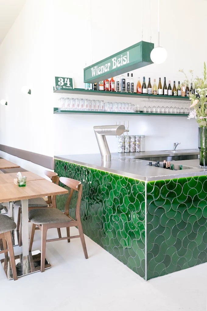 An Austrian restaurant has been redesigned to boast a striking bar counter covered in tessellated emerald green glazed tiles, warm wood furniture, chrome accents, and pendant spherical lighting.