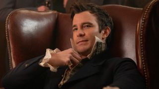 Benedict Bridgerton slouching in a chair and holding his button on his collar. 