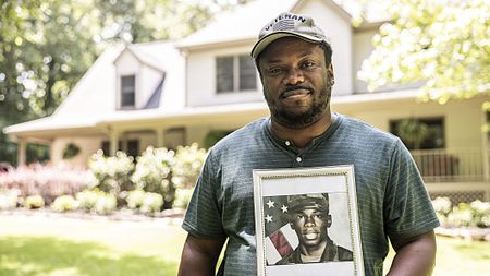 Portrait of a U.S. military veteran holding a framed service portrait of himself in front of his home. He is wearing a cap that says "veteran."