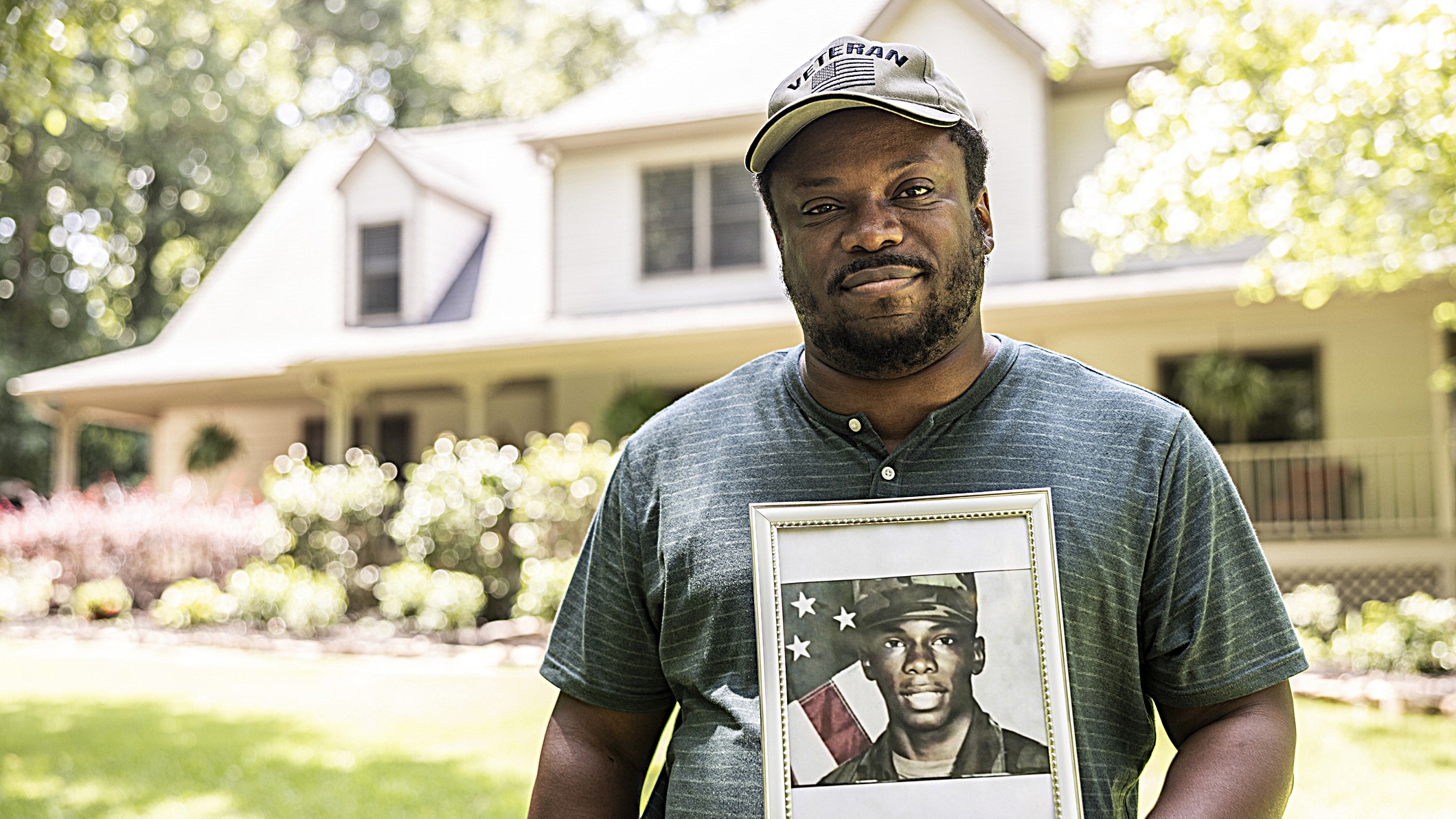 Portrait of a U.S. military veteran holding a framed service portrait of himself in front of his home. He is wearing a cap that says "veteran."