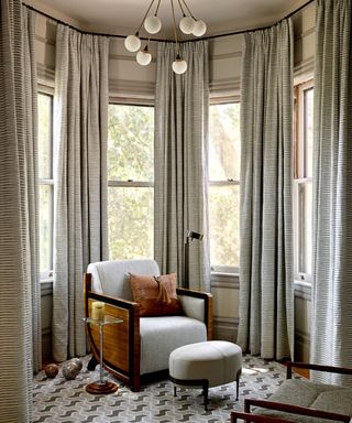 A sitting area in a bay corner with a chair and footstool and floor to ceiling curtains