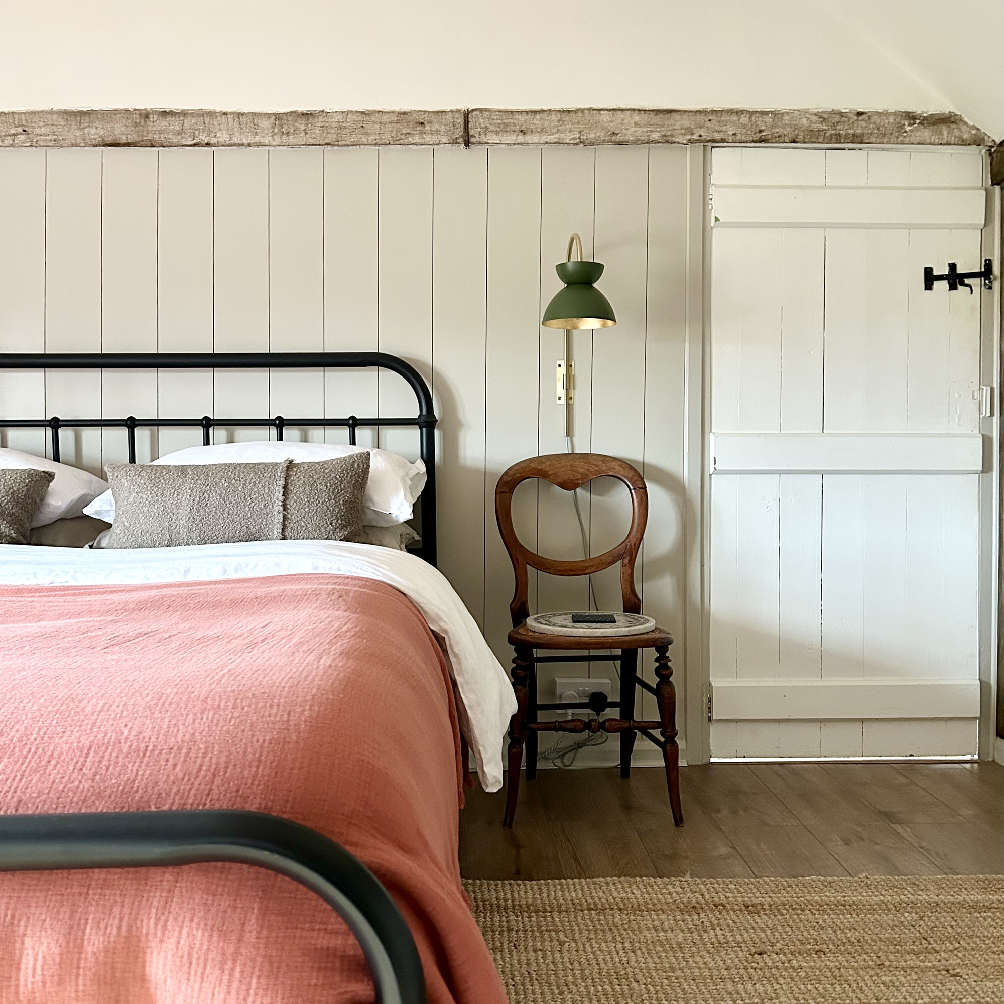Country bedroom with wood panelling and black iron framed bed, dressed with a terracotta linen throw, with a vintage chair used as a bedside table