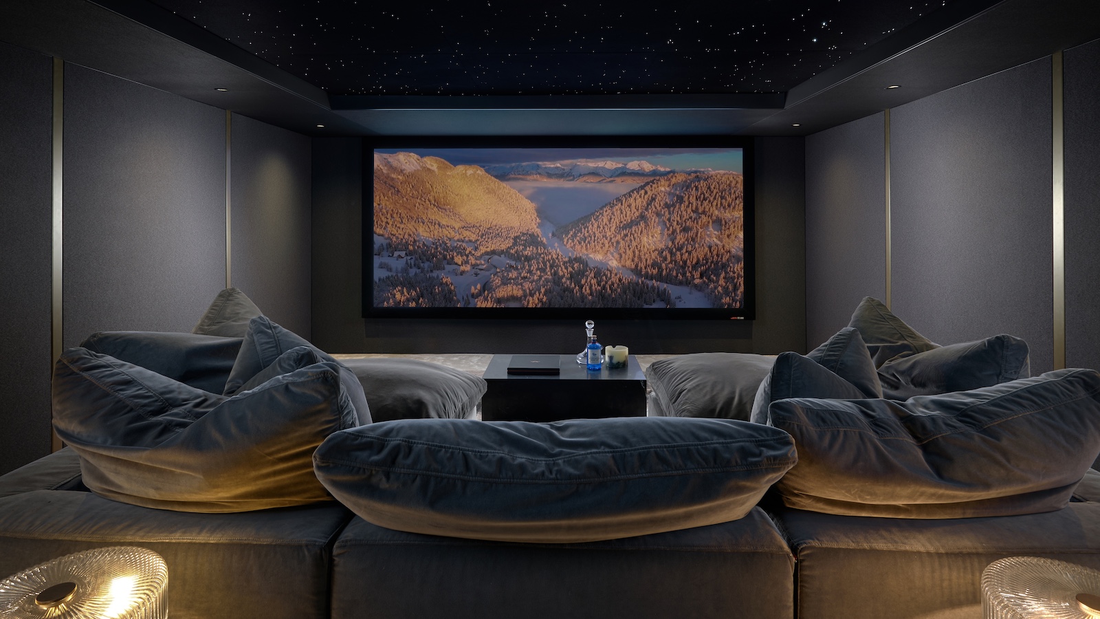 A photograph of a luxury, custom-installed home cinema with a huge projector screen, lush u-shaped sofa, grey walls and lamps
