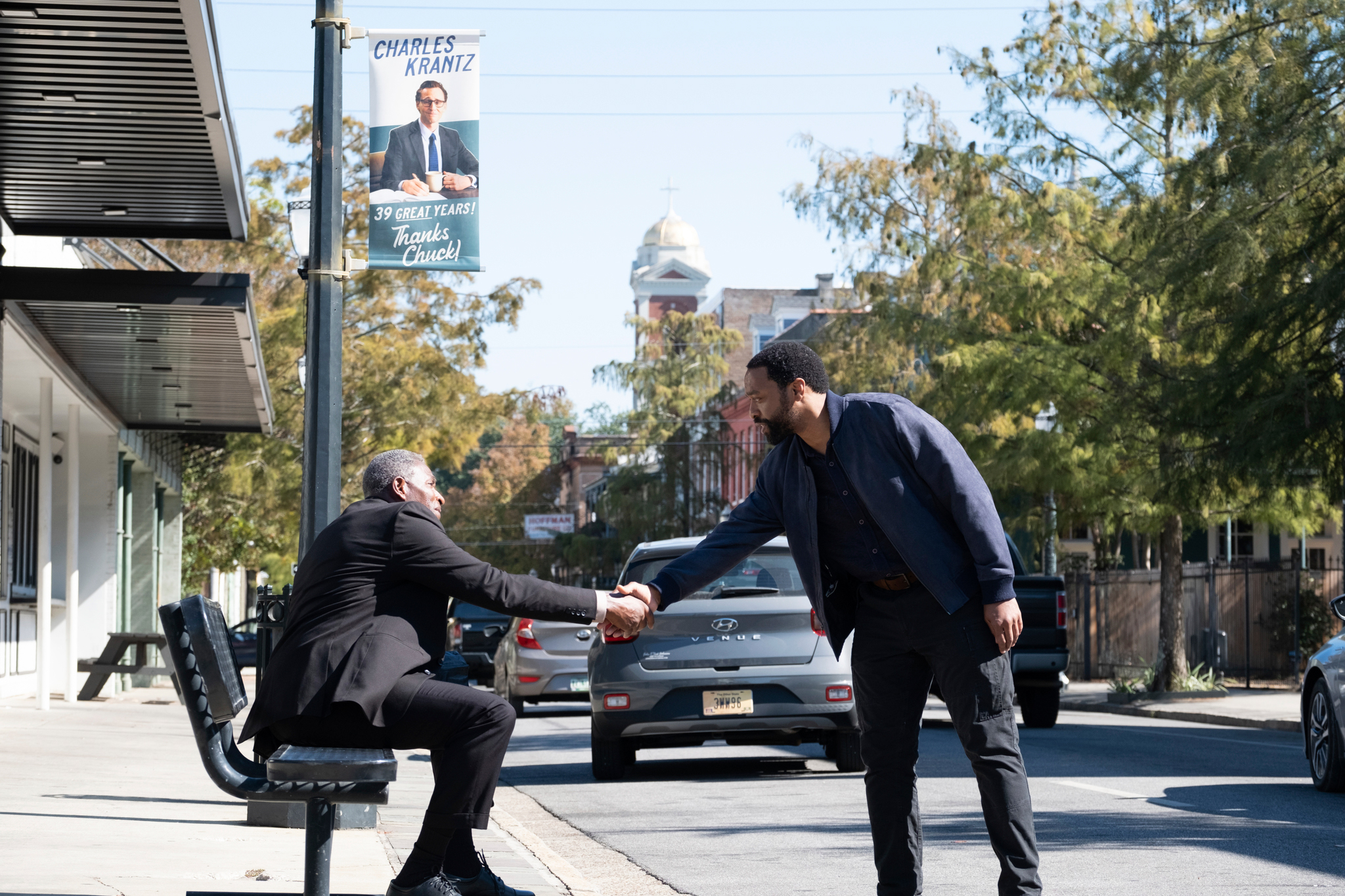 Two men (Carl Lumbly and Chiwetel Ejiofor) shake hands under a sign reading "Charles Krantz. 39 great years! Thanks Chuck!" in 'The Life of Chuck.'
