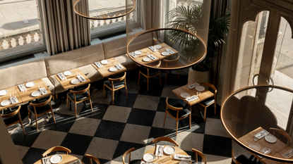An aerial view of a restaurant's dining room, showing checkered, black and white floors, wooden tables with leather and wood chairs in a mid-century design, trailing plants, and circular, pendant chandeliers, all brought to life by airy windows that let plenty of light in.