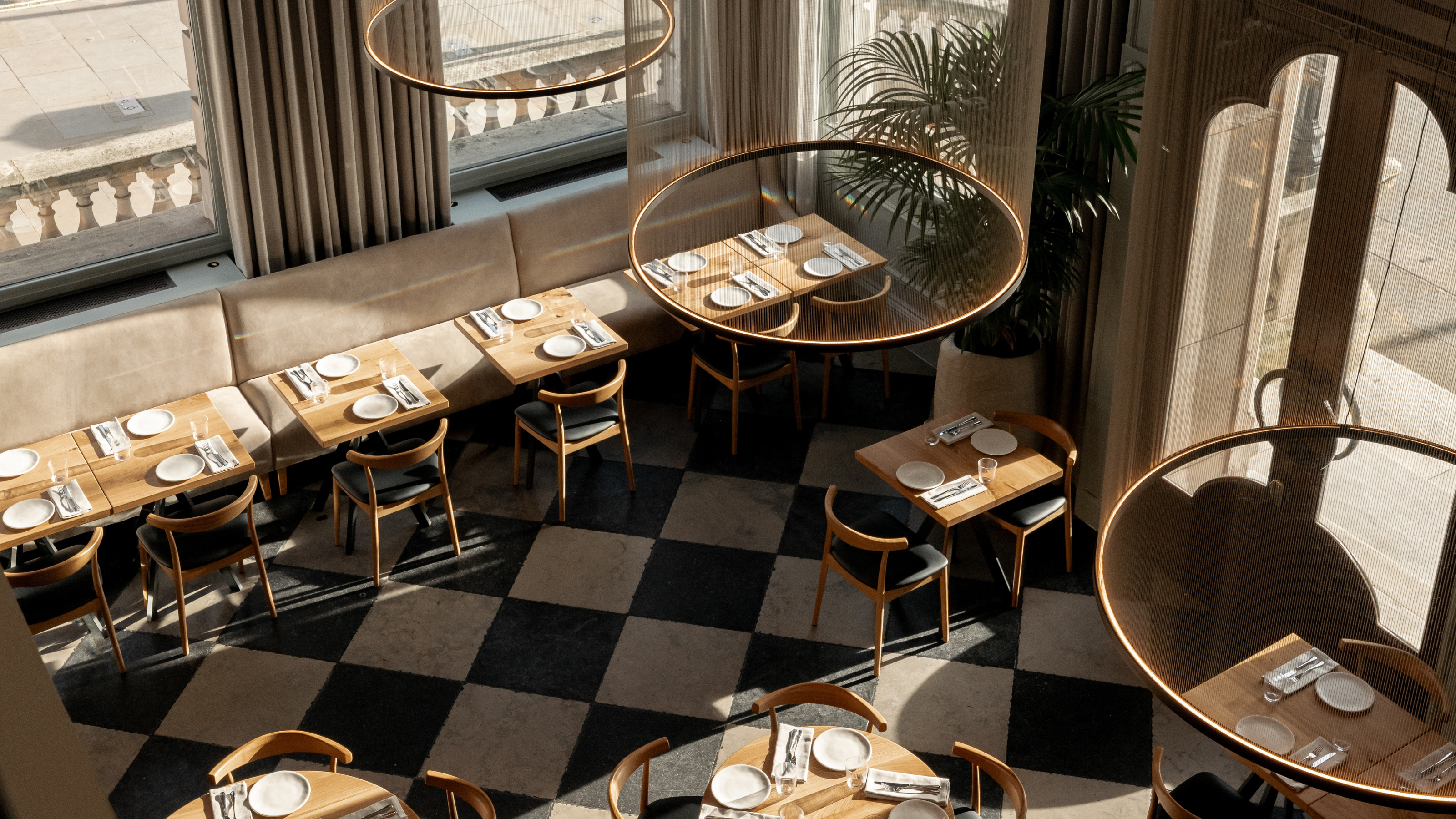An aerial view of a restaurant's dining room, showing checkered, black and white floors, wooden tables with leather and wood chairs in a mid-century design, trailing plants, and circular, pendant chandeliers, all brought to life by airy windows that let plenty of light in.