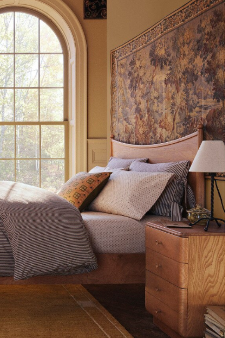 West Elm Pierce &amp;amp; Ward Burl Wood Bedside Table in a contemporary bedroom with a large tapestry behind the bed,large arched windows and a table lamp on top of the bedside with books and magzones on the floor next to it