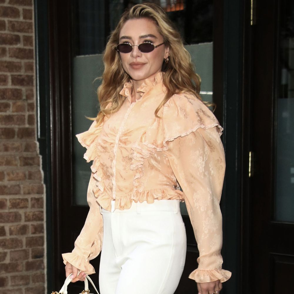 Florence Pugh steps out in New York wearing white flared trousers over heels. She styles her trousers in a peach frill blouse and carries a woven top handle bag.