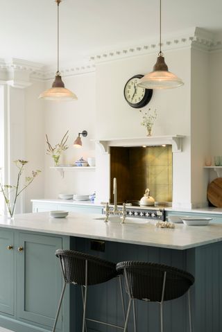 A kitchen with teal blue shaker-style island, two black bar stools, two large glass pendant ceiling lights over island and hob area with metallic gold detail