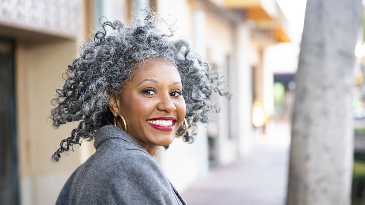 Black woman with grey curly hair and red lipstick wears grey coat and gold hoop earrings