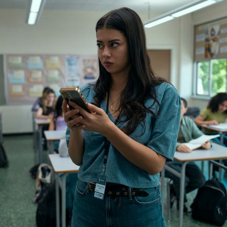 A woman in a blue shirt (Amber Midthunder as Rita Desjardin) uses her phone while standing at the front of a high-school classroom, in Prime Video's 'Carrie.'