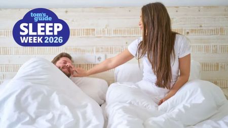 The image shows a woman with brown hair and a white t shirt sitting up in bed with a white duvet pulled up to her waist and white pillow behind her. She leans one arm towards a man lying in bed next to her, whom she is facing. Overlaid in the top left hand corner a blue cloud-shaped badge reads 'Tom's Guide Sleep Week 2026;