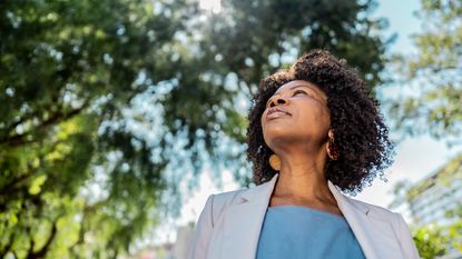 A Gen X woman stands outside under a tree and looks into the distance, appearing thoughtful.