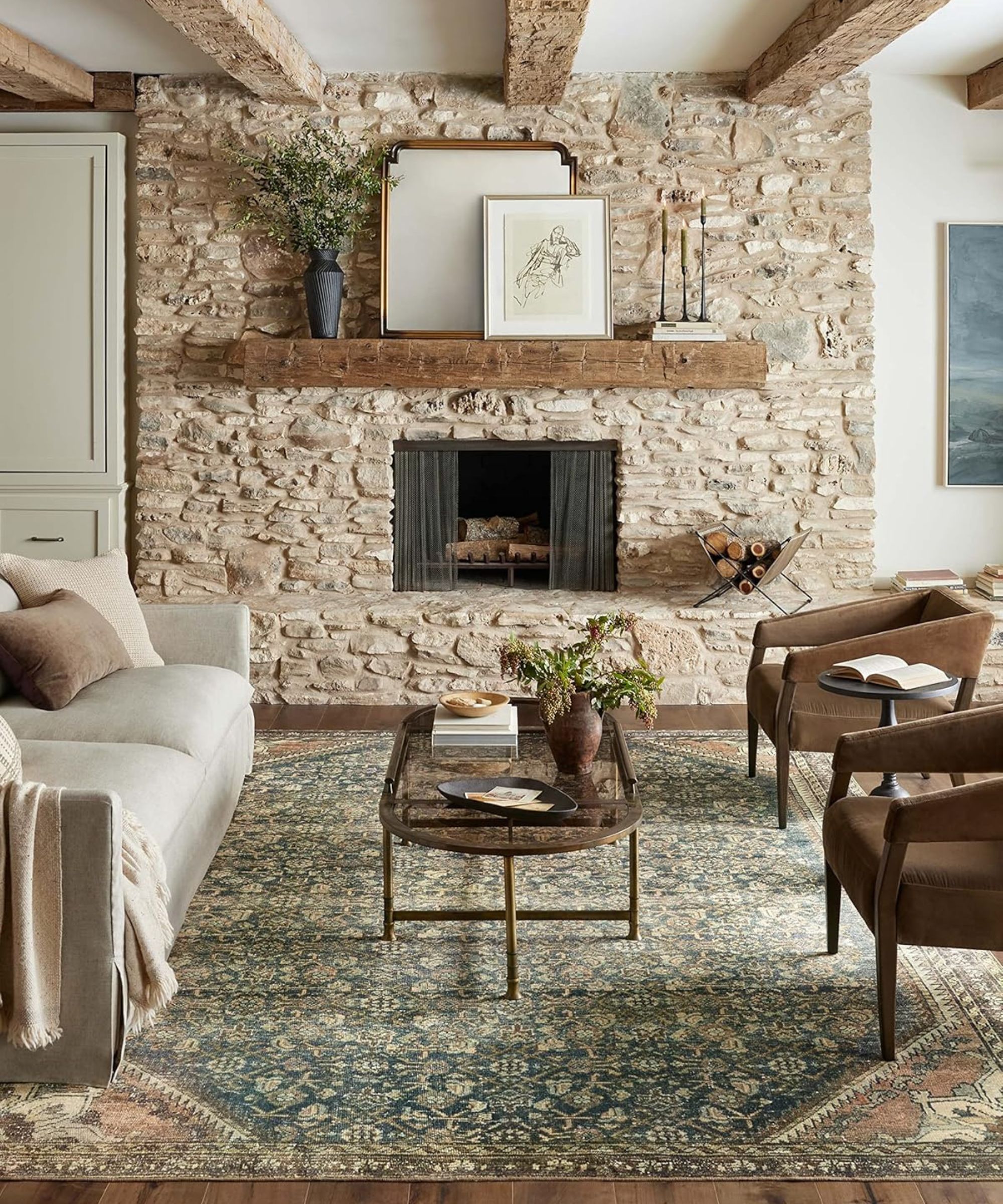 A living room with a stone fireplace, wooden shelf above and a large Persian-style rug in the middle
