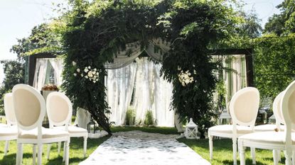 an outdoor wedding set-up, with white chairs, a large foliage arch, and a white carpet