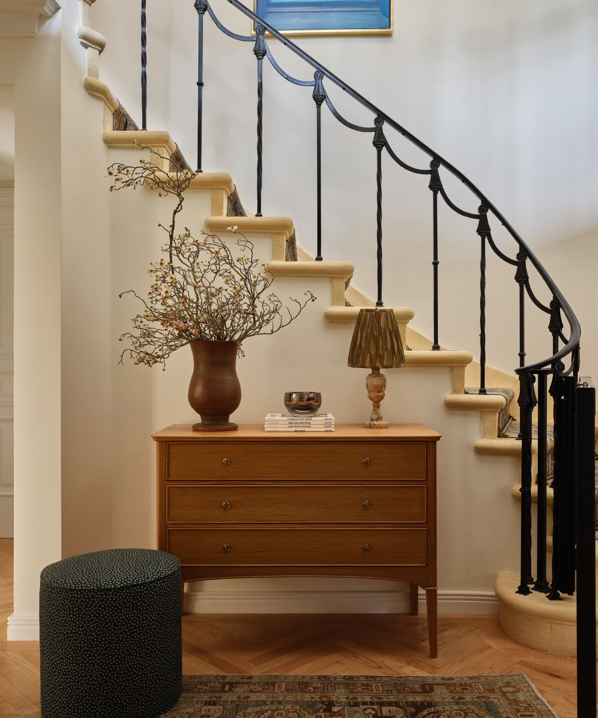 Entryway featuring a curved staircase with a black wrought-iron railing, a mid-century wooden three-drawer dresser, a decorative lamp, and a tall vase with dried branches