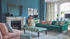 A blue living room with a large white mantelpiece with a mirror above, a green sofa, cushions, a green and white ottoman, a pink sofa with cushions, and a patterned stool. To the right are large bay windows with white curtains.