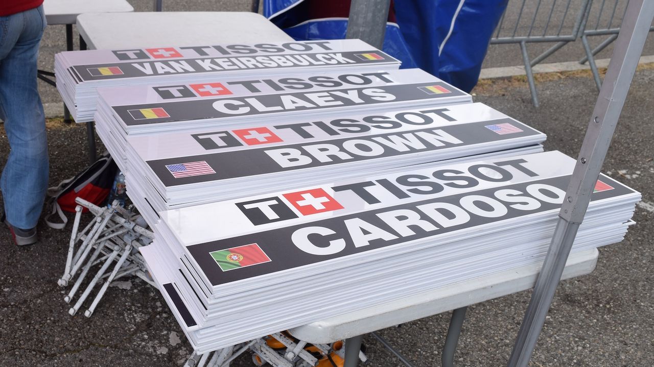 Each rider has a dedicated team car following them, marked with their name ahead of the start