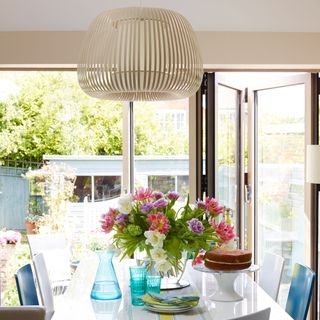 White dining table, flowers in vase, wood pendant light, blue and white chairs and bifold doors