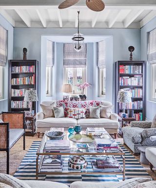 Living room with blue walls, white ceiling beams, bay window, upholstered armchairs and sofa, central glass coffee table.