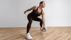 Antonietta Vicario exercises in a studio. She is in a squat position, holding a dumbbell in her left hand near the floor, while her right arm extends behind. 