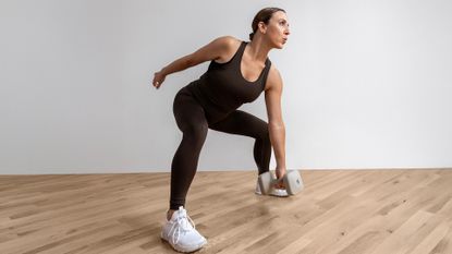Antonietta Vicario exercises in a studio. She is in a squat position, holding a dumbbell in her left hand near the floor, while her right arm extends behind.