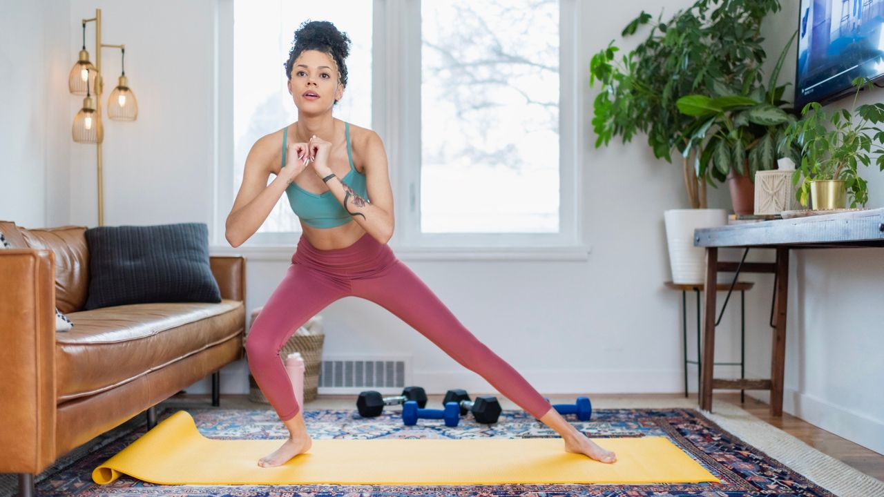 woman wearing pink leggings and turquoise crop top doing a side lunge on a yellow exercise mag in a living room setting. 