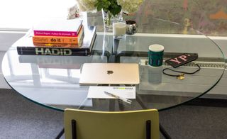 Pale lime green chair and round glass desk with books, an Apple MacBook laptop, a mobile phone, a candle and white flowers with green foliage