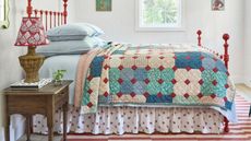 a cottage style bedroom with a rug striped rug, red wrought iron bed, blue linens, a floral bed skirt and patchwork quilt with a wooden nightstand and rattan lamp