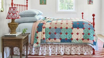 a cottage style bedroom with a rug striped rug, red wrought iron bed, blue linens, a floral bed skirt and patchwork quilt with a wooden nightstand and rattan lamp