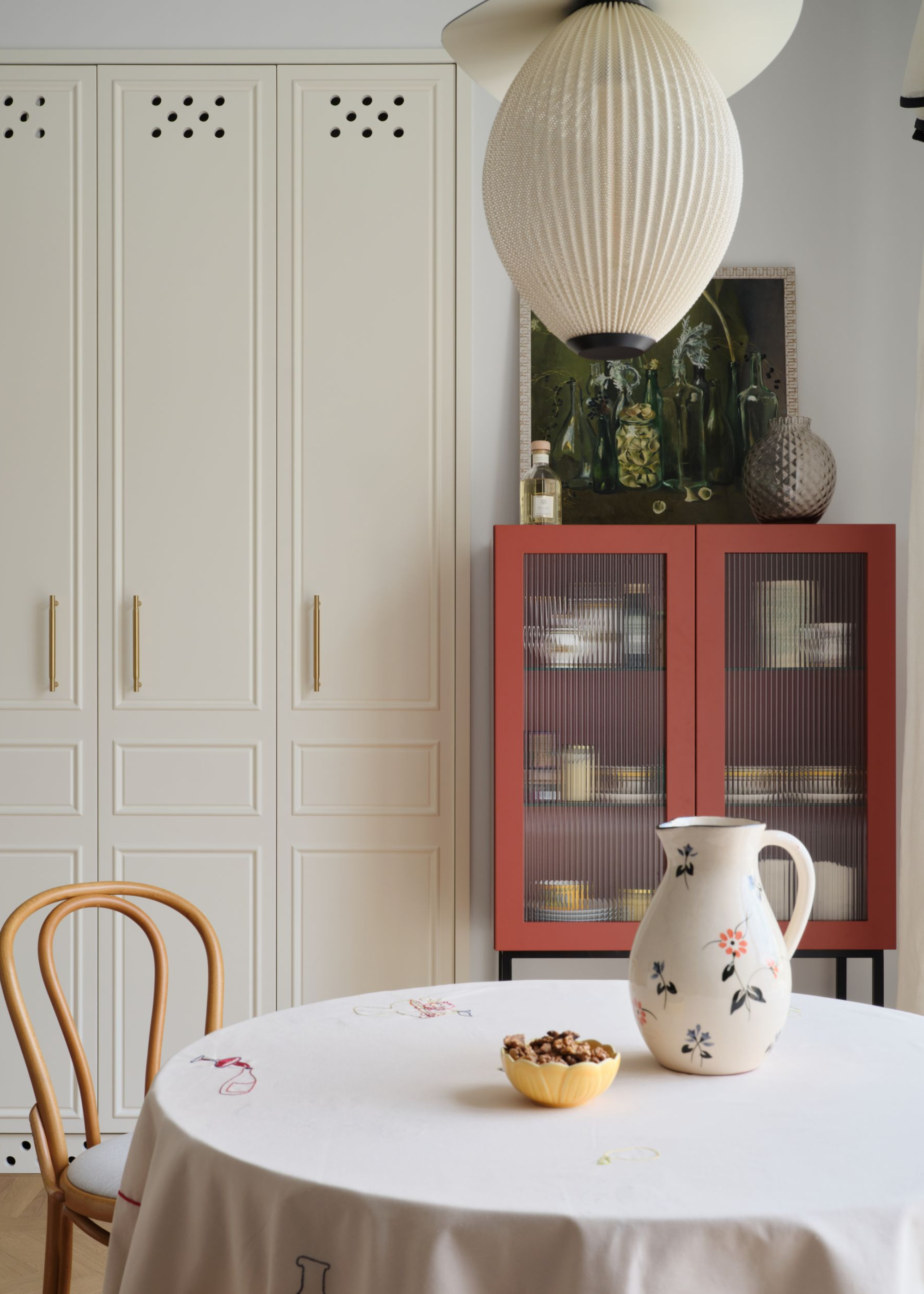 small dining table with white tablecloth and a jug, paper lantern above, and a reeded glass cabinet behind