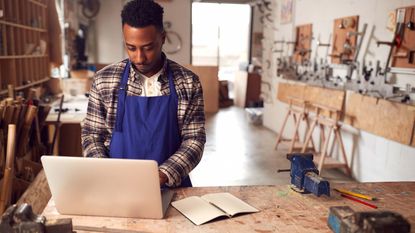 A craftsman looks at his laptop in a his carpentry workshop.