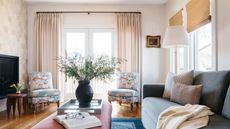 Living room with tiled wall, balcony, wooden floor, pink coffee table with tray and vase with foliage, floral armchairs and rattan blinds on windows