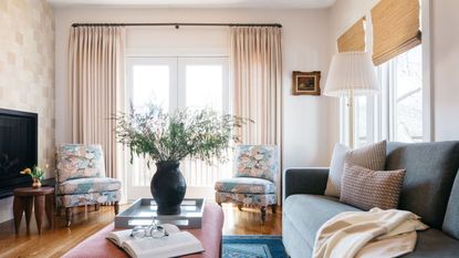 Living room with tiled wall, balcony, wooden floor, pink coffee table with tray and vase with foliage, floral armchairs and rattan blinds on windows