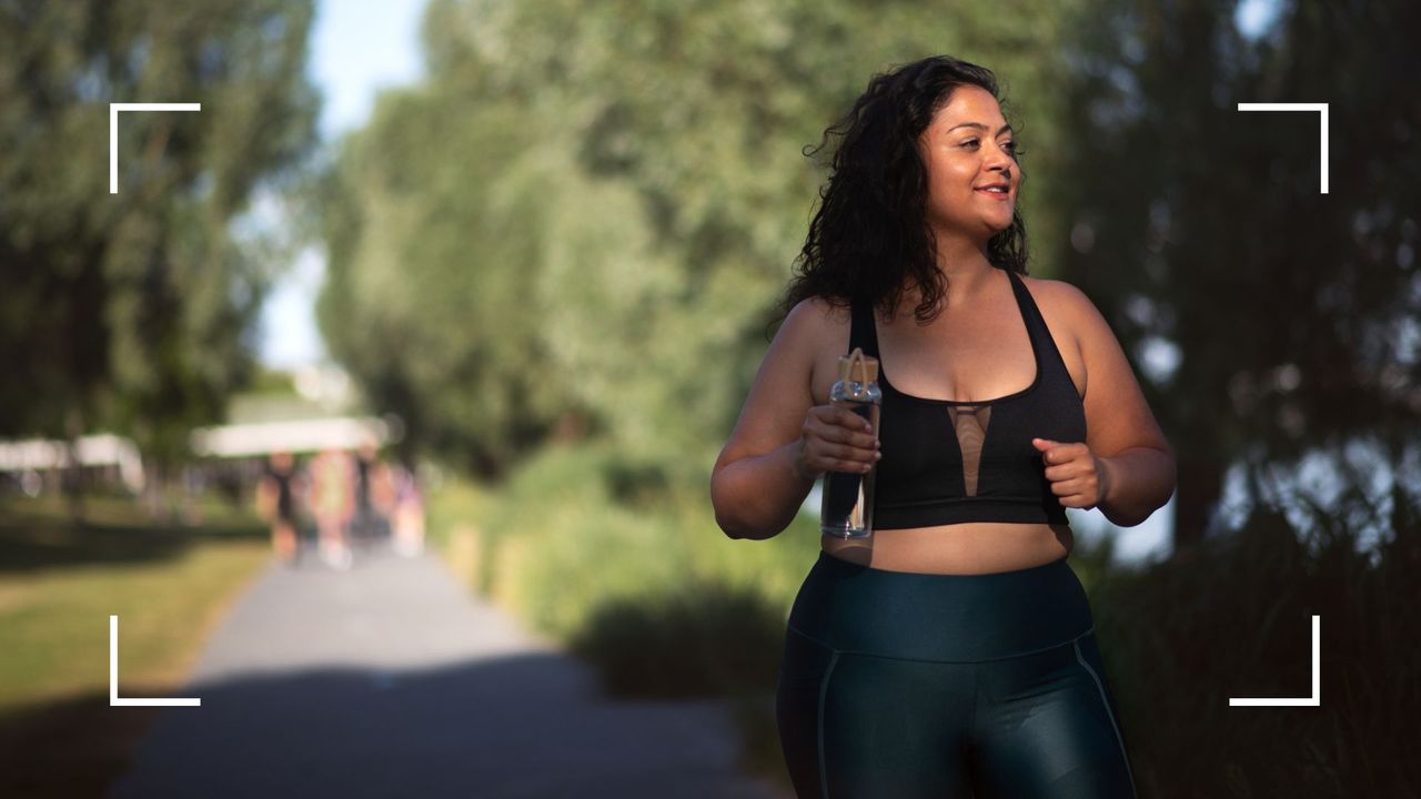 Woman walking through green open space in sports bra and leggings, carrying water bottle, after wondering do you burn more calories in the heat