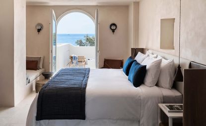 A hotel room with a bed, wooden chair, wooden side table, wooden headboard, round coffee tables and an arched doorway onto a balcony with an ocean view.