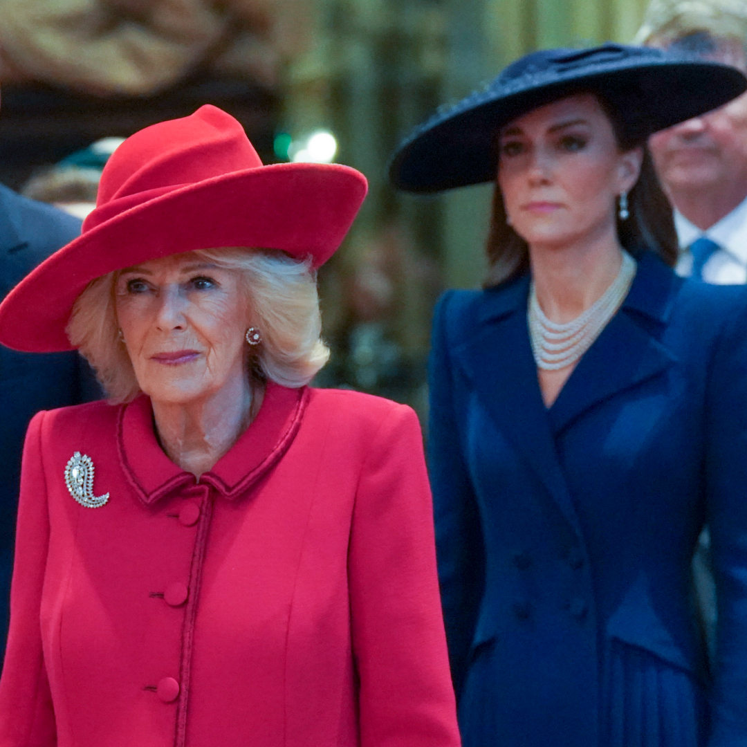 Queen Camilla wears a red coat dress and matching hat while Princess Kate stands behind her wearing a navy coat and hat