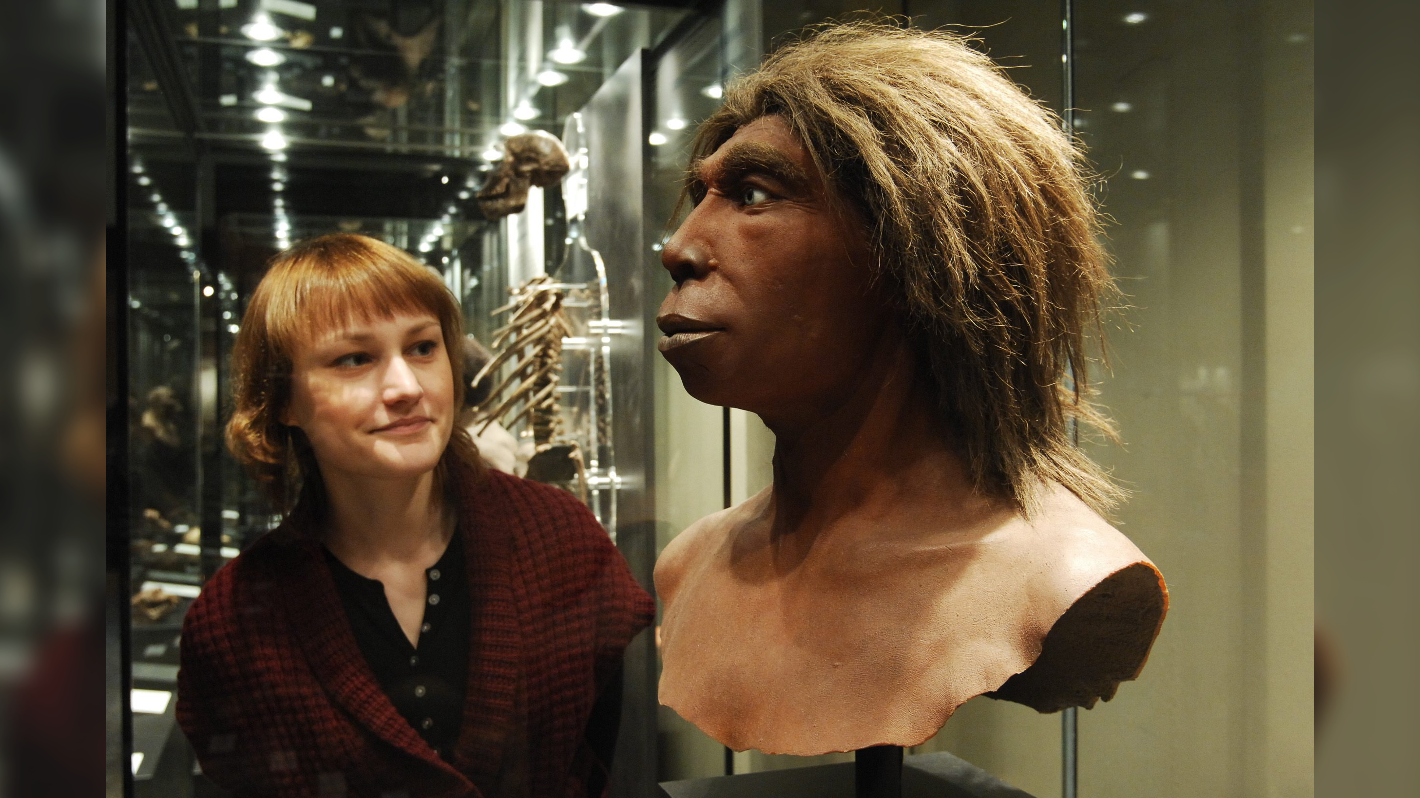 a light-skinned woman with red hair looks at a bust ofa. Neanderthal man in a museum