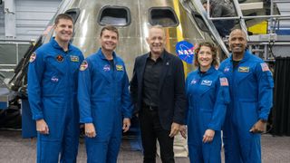 tom hanks with four astronauts, two on either side. all are standing and smiling. the astronauts wear blue flight suits. tom hanks is in black. behind is a cone-shaped spacecraft simulator with oval windows