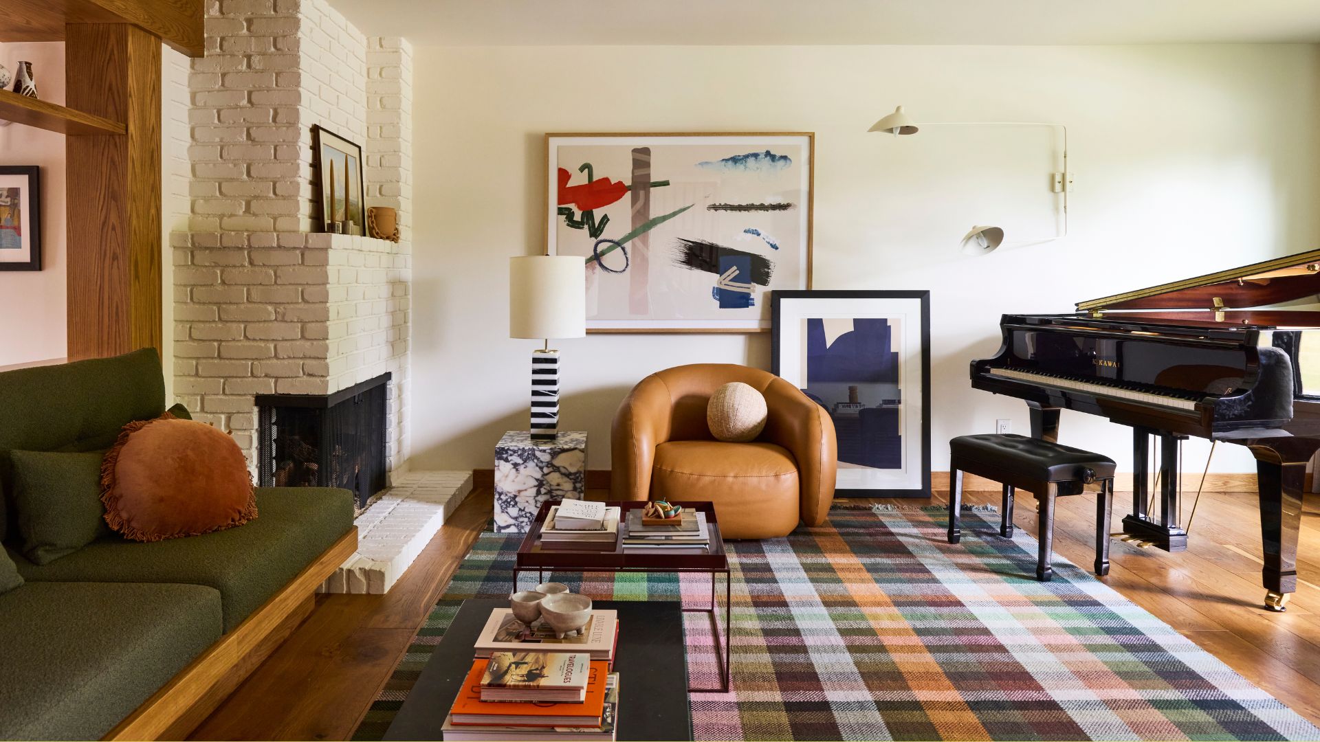 A white living room with a brick fireplace, a painting on the wall and leaning below, a white extending wall sconce, a brown leather armchair with a round cushion, a stone pedestal with a  striped lamp, a colorful checked rug, a black coffee table with books, a green couch with a mustard cushion, and a grand piano with a bench