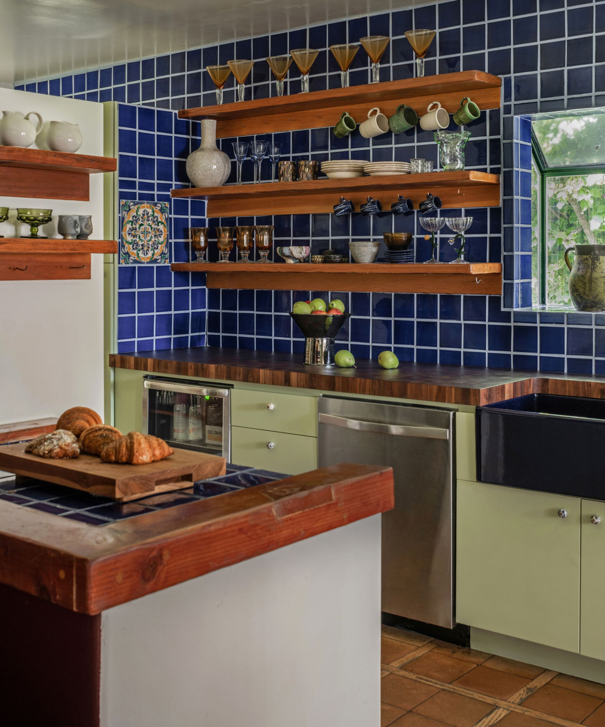 A modern, small kitchen with pistachio green lower cabinets, electric blue wall tiles, open shelving, and terracotta flooring.