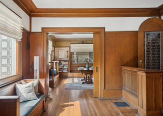 An interior photo of a room with wood panelled walls, a window seat, and hard wood flooring with a view through an open doorway