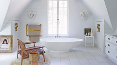an all white attic bathroom with a freestanding tub in the centre with a brown armchair sat next to it