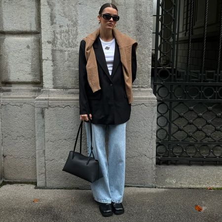 Mary wears a white t-shirt, black blazer, brown sweater, blue jeans, black boots, and a large black east-west bag.