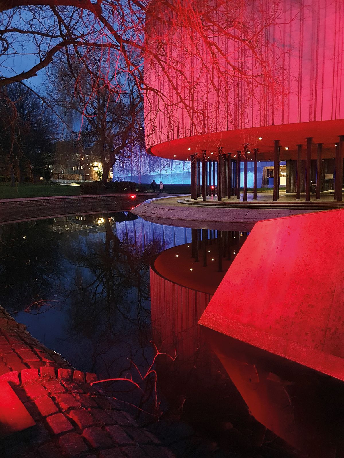 A modern building glows red at dusk, reflected in a calm pond, surrounded by trees and people strolling nearby