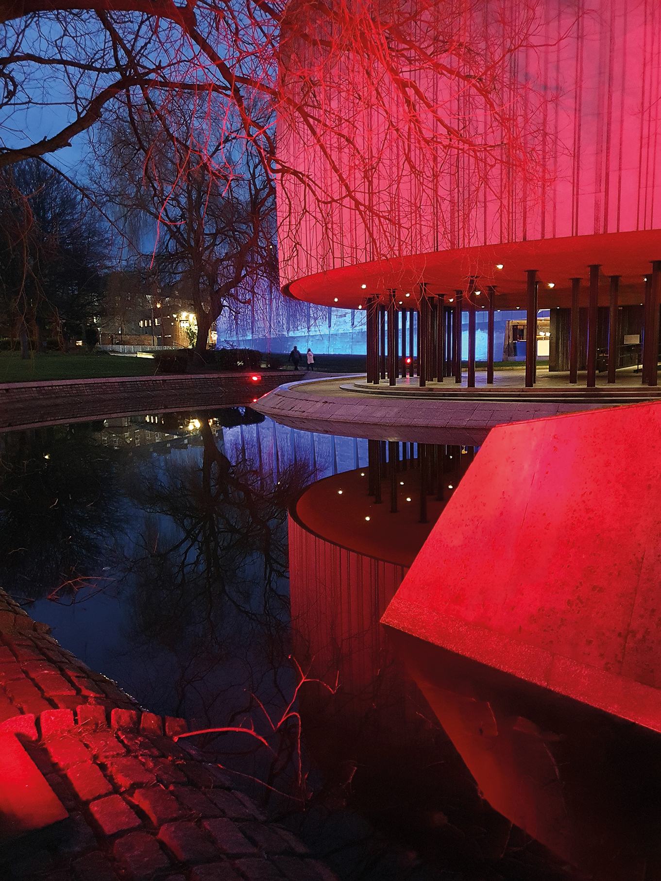 A modern building glows red at dusk, reflected in a calm pond, surrounded by trees and people strolling nearby
