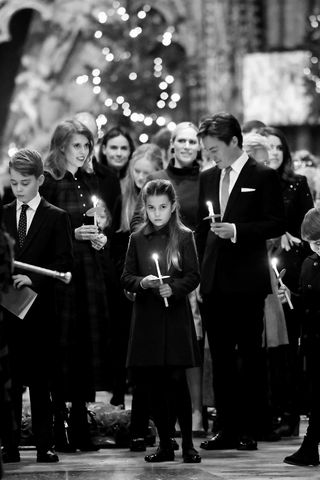 A black and white photo of Princess Charlotte standing in the middle of Westminster Abbey holding a lit candle and surrounded by Pricne George, Princess Beatrice, Edo Mapelli Mozzi, Zara Tindall and Sophie Winkleman