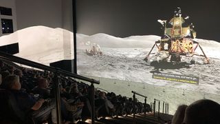 a panoramic photo of an Apollo lunar module on the moon is projected on a giant movie screen, as well as the theater's walls and floor