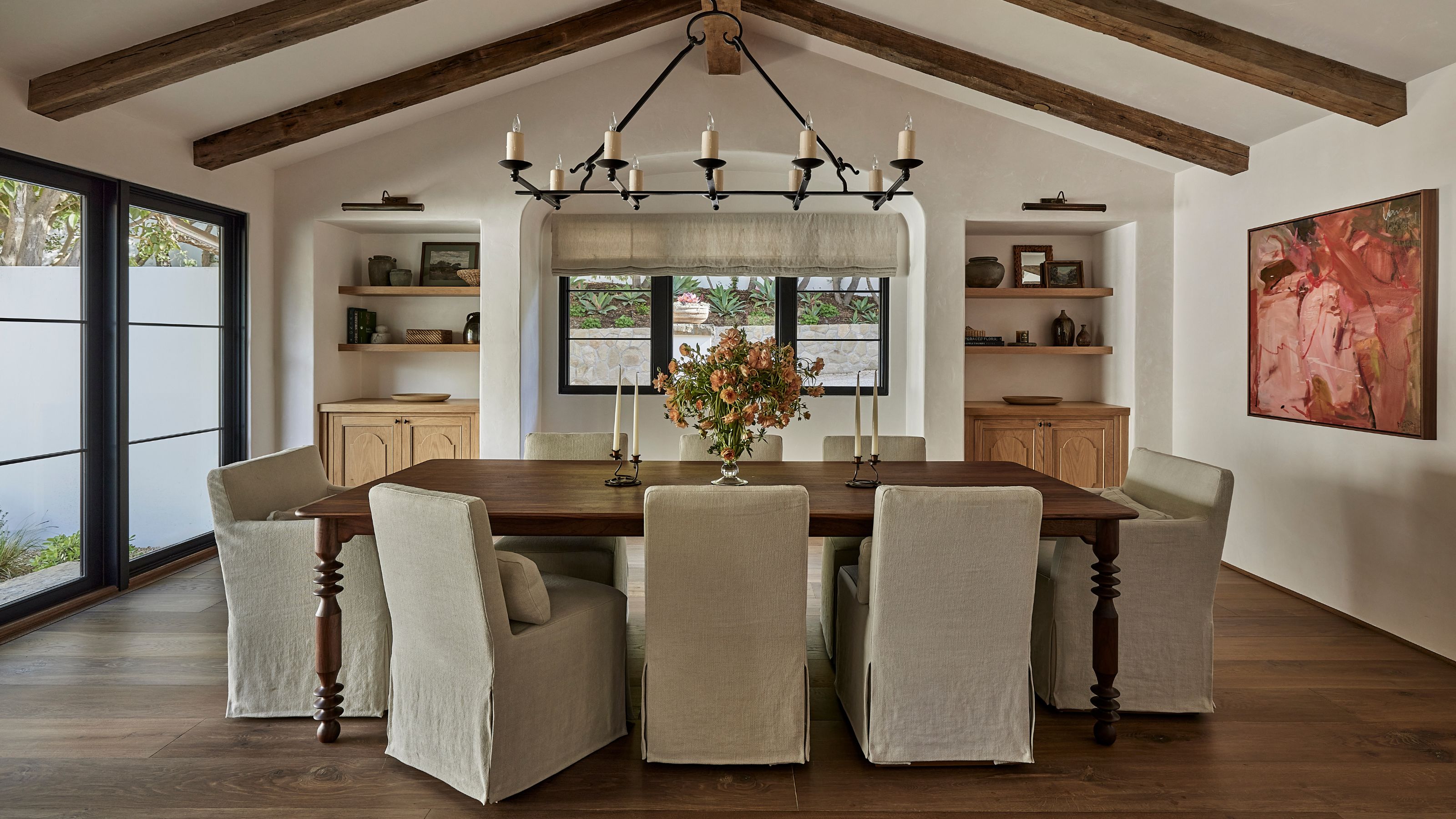 a vaulted dining room with a beamed ceiling, open shelved alcoves, large patio doors and a wooden table with linen slipcovered chairs